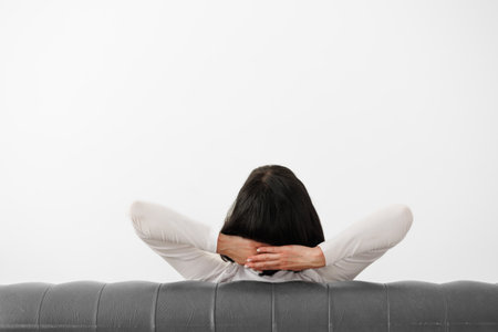 Rear view at relaxed woman with hands behind head sits on comfortable leather sofa on white wall background, feels mental balance, dreams of satisfaction, enjoys no stress free peaceful. Copy spaceの写真素材
