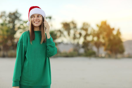 Happy young woman in Santa Claus Christmas red hat and green sweater talks with smile on mobile cell...
