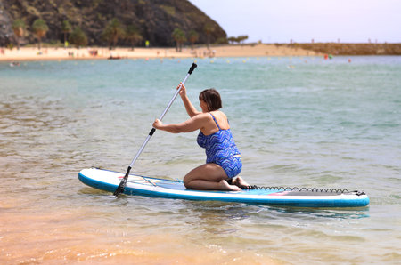 Back view of adult woman in swimsuit sits on inflatable SUP board and paddles through sea...
