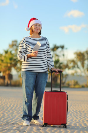 Independent mature woman in Christmas Santa Claus hat, striped shirt holds passport, boarding ticket, suitcase on seashore with palms backdrop celebrates new year holidays vacation at exotic resort.の写真素材