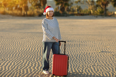 Happy independent adult woman 50-55s in Christmas Santa Claus hat, striped shirt holds red suitcase on sand seashore with palms backdrop celebrates new year holidays vacation alone at tropical resort.の写真素材