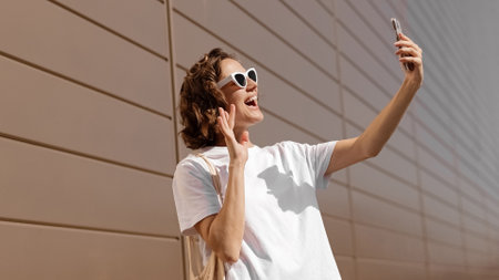 Happy young woman in stylish sunglasses taking selfie photo on smartphone on sunny summer day. Female blogger uses mobile phone camera, attractive caucasian girl touching hair for selfie pictureの写真素材