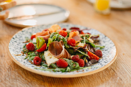 Close up of served fresh salad with jamon, raspberries, tomatoes cherry, cheese and mixed lettuce on plate, dressed olive oil, balsamic vinegar and spices on wooden table at restaurant. Food at eventの写真素材
