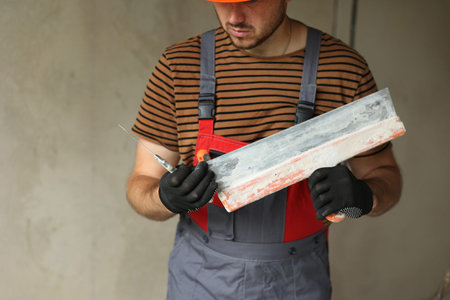 Worker in overalls and safety hard hat holds and prepares two spatulas for plastering concrete wall...