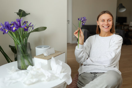 Allergy free. Beautiful young woman holds and sniffs iris flower, enjoys of smell without of runny nose symptoms at home. Bouquet of spring flowers at vase, used napkins on table. Allergic rhinitis.の写真素材