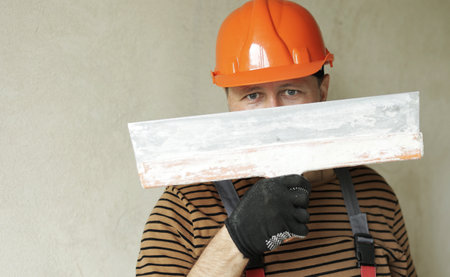 Smiling handyman repairman worker in overalls and protective orange helmet holds big putty knife for plastering concrete wall with putty. Renovation at apartment and construction new building conceptの写真素材