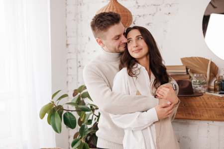 Happy young couple in love is hugging gentle and smiling together celebrating Valentines day at cozy Scandinavian style home. Man and woman enjoying spending time together, relaxing on dateの写真素材