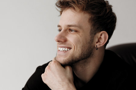 Cheerful young handsome hipster tattooed man 20-25 years in casual clothes smiles with teeth indoor. Emotional caucasian guy with stylish haircut, black earring in ear holds hand at chin, looks side.の写真素材