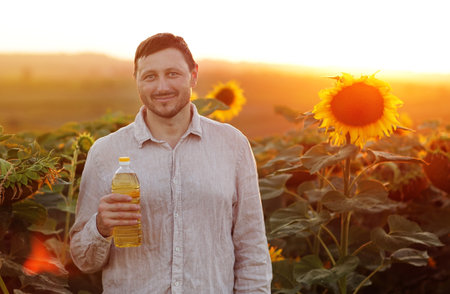 Male farmer holds plastic bottle of sunflower oil at blooming sunflower field at sunset. Smiling businessman agronomist checks and controls of harvest quality. Organic agriculture and agribusiness.の写真素材