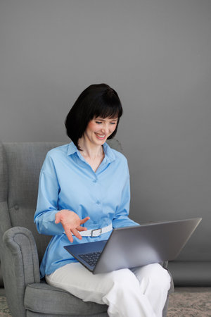 Positive adult business woman mentor teacher psychologist sits on armchair consulates talk in online seminar on laptop,work with patients at home in remote, teach advice by digital training at office.の写真素材