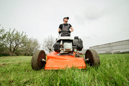 Professional gardener in protective apparel is mowing green grass lawn using modern gasoline...