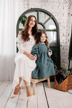 Mothers day. Elegant young mom and little cute daughter sit and smile, embrace on wooden chair at living room at home, look at camera. Trust, support and love between mommy and child. Children day.の写真素材