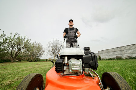 Professional gardener in protective apparel is mowing green grass lawn using modern gasoline cordless lawnmower at backyard. Seasonal landscaping design work. Blooming trees on background.の写真素材