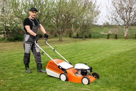 Professional gardener in protective apparel is mowing green grass lawn using modern gasoline cordless lawnmower at backyard. Seasonal landscaping design work. Blooming trees on background.の写真素材