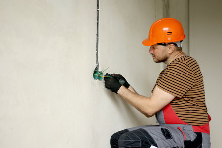 Electrician male in uniform, protective gloves and helmet checks presence of electrical voltage in socket phase uses electrical tester screwdriver.Examining wires in outlet by voltage detector indoor.の写真素材
