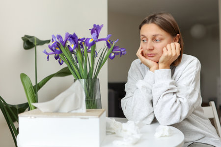 Seasonal allergy. Unhealthy allergic young woman uses paper tissues, suffers from runny nose, snees from flowers pollen at kitchen home by table. Girl with flu, itching or cough, allergy rhinitis.の写真素材