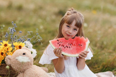 Adorable little child girl bites big slice of watermelon at picnic with teddy bear, wild flowers outdoors on sunny day. Healthy fruit snack for children. Summer holidays concept and happy childhoodの写真素材