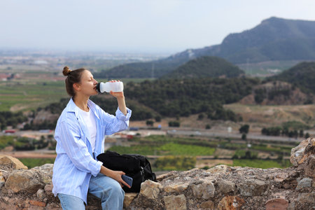 Tired after hiking, young woman traveler with a backpack drinks fresh water from a bottle against the backdrop of a mountains. Adventure, independent travel, tourism, trekking, ecotourism on holidays.の写真素材