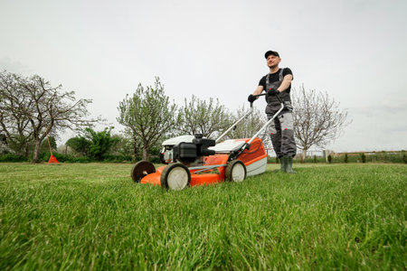 Professional gardener in protective apparel is mowing green grass lawn using modern gasoline cordless lawnmower at backyard. Seasonal landscaping design work. Blooming trees on background.の写真素材