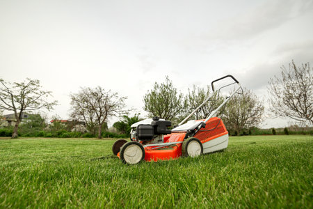 side view of modern orange-gray gasoline lawn mower cutting bright lush green grass. Gardening work tools. Rotary lawn mower machine on lawn. Professional law care service. Place for text.の写真素材