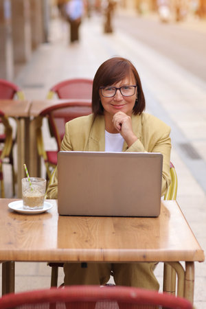 Mature stylish confident businesswoman in eyeglasses and suit works on laptop drinks coffee at European cafe outdoors. Concept of remote work from public place, digital freelance and modern lifestyle.の写真素材