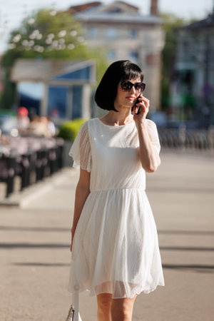 Romantic young brunette woman with short hair wears mini white dress and sunglasses, talking on cellphone, walking on a crowded city street in the summertime. Girl communicates by smartphone outdoors.の写真素材