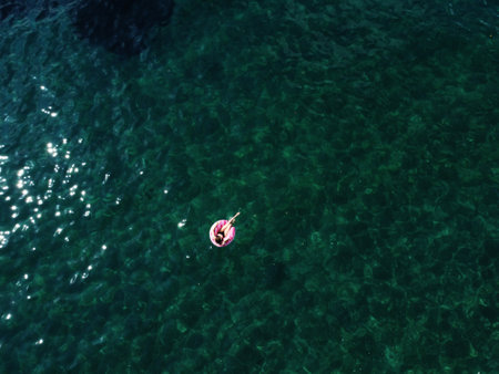 Aerial drone view of young woman with open arms in swimsuit lying and relaxing on big pink donut float swims on water surface with transparent bottom. Summer holiday vacation at exotic resort.の写真素材