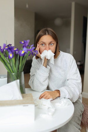 Unhappy sick young woman with paper tissues inside nose from runny nose and sneezes sits by table at home. Female with pollen allergy, flu, cold, rhinitis, sinusitis and with viral infection.の写真素材