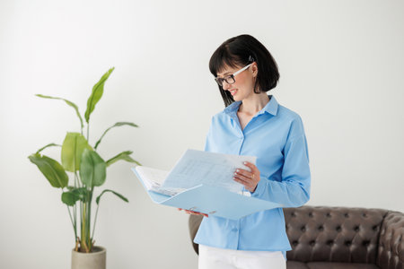 Young businesswoman holds paper documents folder, calculations and analyzes financial reports, reviewing bills on sofa at office with plant. Lawyer, legal adviser, office assistant or company managerの写真素材