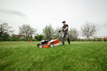 Professional gardener in protective apparel is mowing green grass lawn using modern gasoline cordless lawnmower at backyard. Seasonal landscaping design work. Blooming trees on background.の写真素材