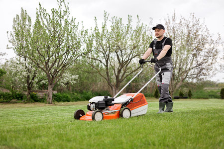 Professional gardener in protective apparel is mowing green grass lawn using modern cordless lawnmower at backyard. Seasonal landscaping design work. Blooming trees on background.の写真素材