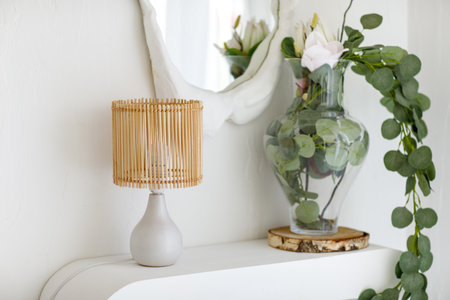 Scandinavian minimalistic interior design with table rattan lamp, eucalyptus branches in glass vase on shelf and mirror with textured white plaster frame on wall in light living room. Cozy home decorの写真素材