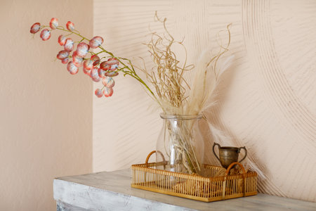 Creative arrangement of artificial branches with red petals, dried grass and feathers in glass vase stands in rattan tray on concrete shelf, beige plastered textured walls. Stylish interior designの写真素材