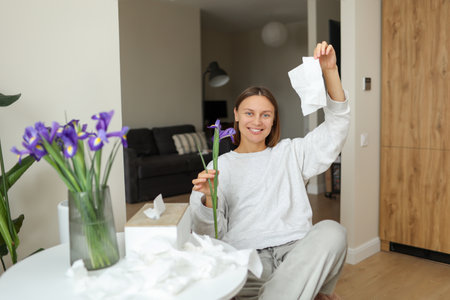 Allergy free. Cheerful young woman throws napkins up at home. Happy life without runny nose, itching or cough symptoms. Allergen bouquet of iris flowers at vase, used paper handkerchiefs on table.の写真素材