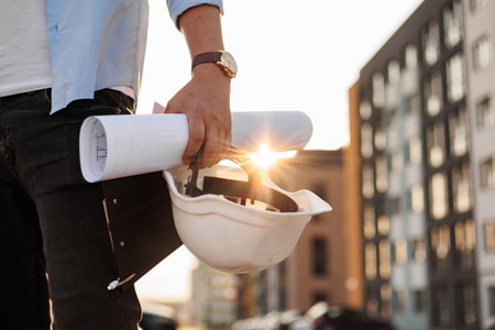 Unrecognisable engineer, architect or worker holds in hand protective white hard hat and blueprint on multi-apartment residential complex background, sunlight. Concept of new real estateの写真素材