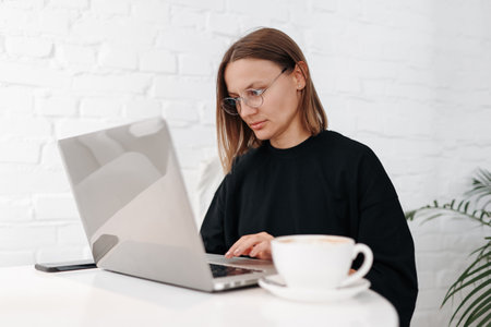 Smiling young woman freelancer brows internet on laptop while drinks coffee at workplace. Female entrepreneur, businesswoman or manager distance works online watches webinars, student studies remote.の写真素材