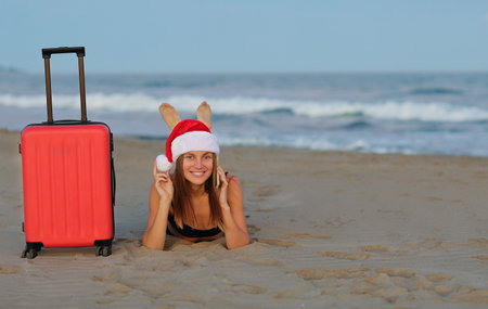 Christmas holidays. Happy young woman in red Santa Claus hat with suitcase lies on sand seashore, books online ticket on smartphone looks at camera.の写真素材