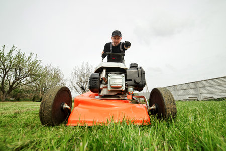 Professional gardener in protective apparel is mowing green grass lawn using modern gasoline cordless lawnmower at backyard. Seasonal landscaping design work. Blooming trees on background.の写真素材