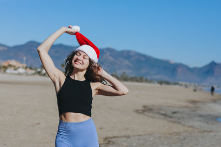 Cheerful young slim woman in Santa hat and sportswear on sand beach and mountains backdrop. Happy New Year 2026, Christmas celebration holiday abroad at hot country. Activity at winter vacationの写真素材