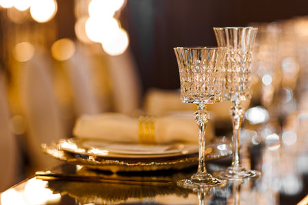 Luxury table setting with plates, napkin with ring, crystal wineglasses and golden cutlery on mirror table for romantic dinner in restaurant, bokeh. Christmas, New Year 2026 dinner. Wedding, birthdayの写真素材