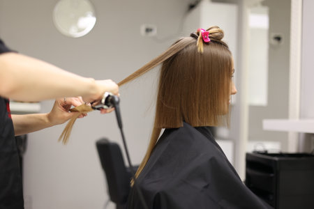 Unrecognisable professional hairdresser ironing female client hair with hair straightener and comb, making stylish hairstyle in beauty salon. Keratin, lamination, hair care, hair salon and hairstyling.の写真素材