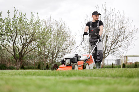 Professional gardener in protective apparel is mowing green grass lawn using modern cordless lawnmower at backyard. Seasonal landscaping design work. Blooming trees on background.の写真素材
