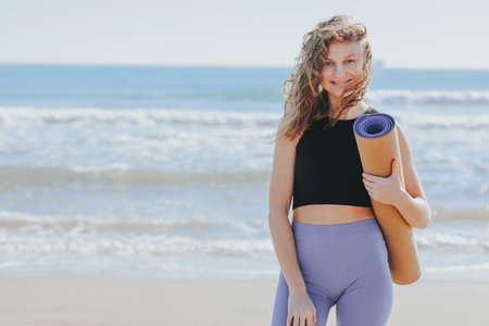 Young woman holds yoga mat, looks at camera and smiles. Lady prepares for workout outdoors. Sport and fitness on seashore. Healthy lifestyle, morning exercises, female wellness and power, healthcare.の写真素材