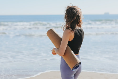 Rear view of slim lady in sportswear holds yoga mat and prepares to workout on sea beach. Sport fitness outdoors. Healthy lifestyle, morning exercise, female wellness and power, healthcare, activitiesの写真素材