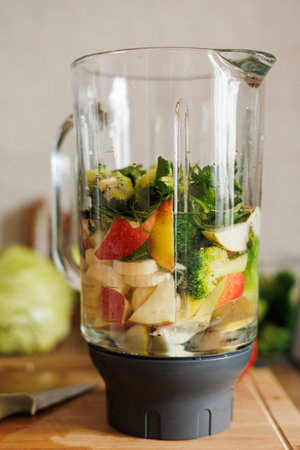 Glass blender bowl with apple, banana and broccoli slices, parsley and water inside on a wooden table on home kitchen. Healthy detox smoothie drink and lifestyle diet. Pieces of fruit ingredients.の写真素材