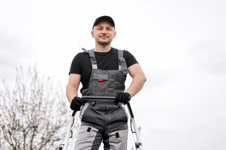 Bottom view of professional male gardener in protective apparel, gloves, cap and overalls holds handle of gasoline cordless lawnmower for mowing grass lawn outdoors. Seasonal landscaping design workの写真素材