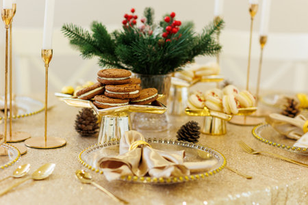 Luxury Christmas dinner table setting. Fir branches in vase, chocolate cookies, macaroons, golden napkin on glass plate, cutlery, candlesticks on tablecloth. Xmas holidays. Festive new 2026 year partyの写真素材