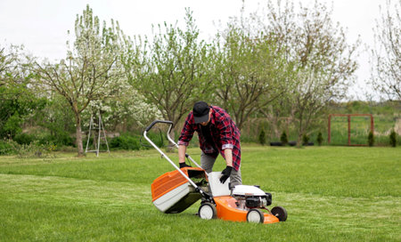 Man in casual clothes takes out bag with cut grass of lawn mower after mowing lawn with at own...