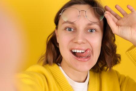 Playful young woman takes a selfie, smiles, sticks out her tongue, holds glasses on yellow backdrop. Modern lifestyle, social media, youth influencer marketing, self expression, personal brandingの写真素材