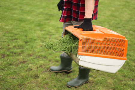 Unrecognisable man in casual clothes mows lawn with lawn mower at own backyard, holds bag with cut...